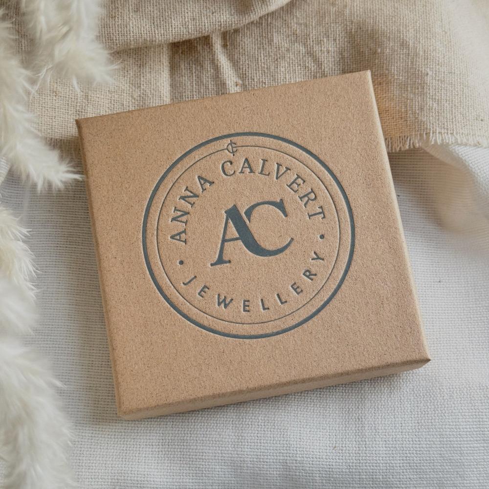 A brown cardboard jewellery box with 'ANNA CALVERT JEWELLERY' branding on a cream fabric background