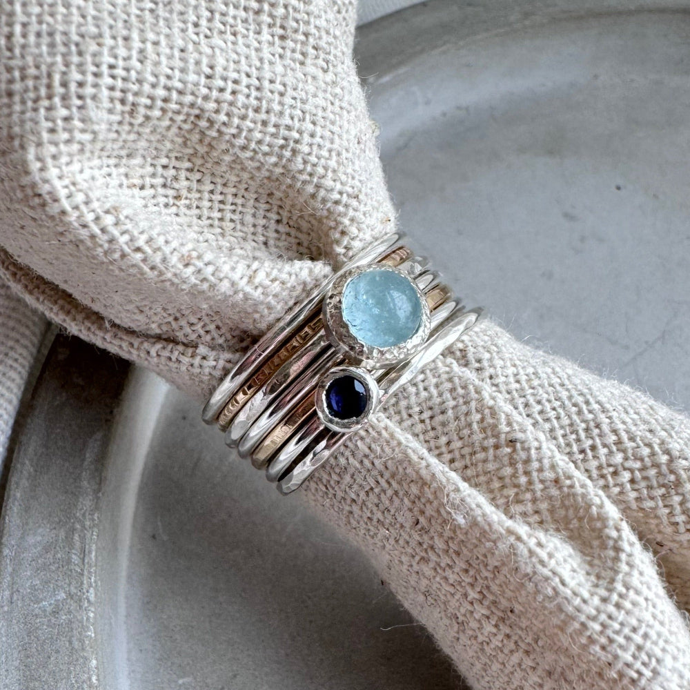 a stack of rings displayed on cream fabric the rings are silver or gold stacking rings with a aquamarine one in the middle and an iolite one next to it 