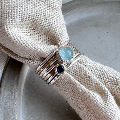a stack of rings displayed on cream fabric the rings are silver or gold stacking rings with a aquamarine one in the middle and an iolite one next to it 