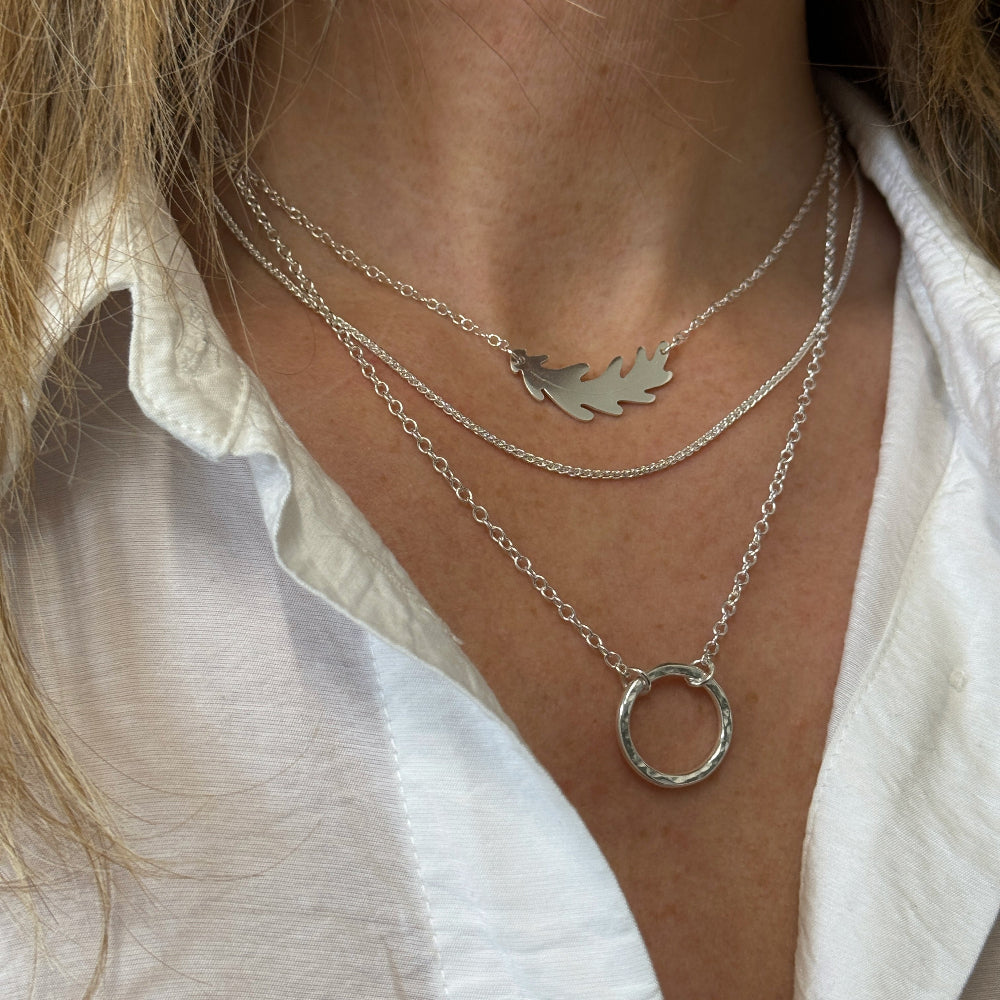 three silver necklaces worn layered. Shortest is an oakl leaf, then a fine chain and the longest is a silver circle pendant