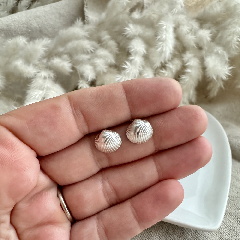 Two small cockle shell earrings held between fingers against a textured fabric background