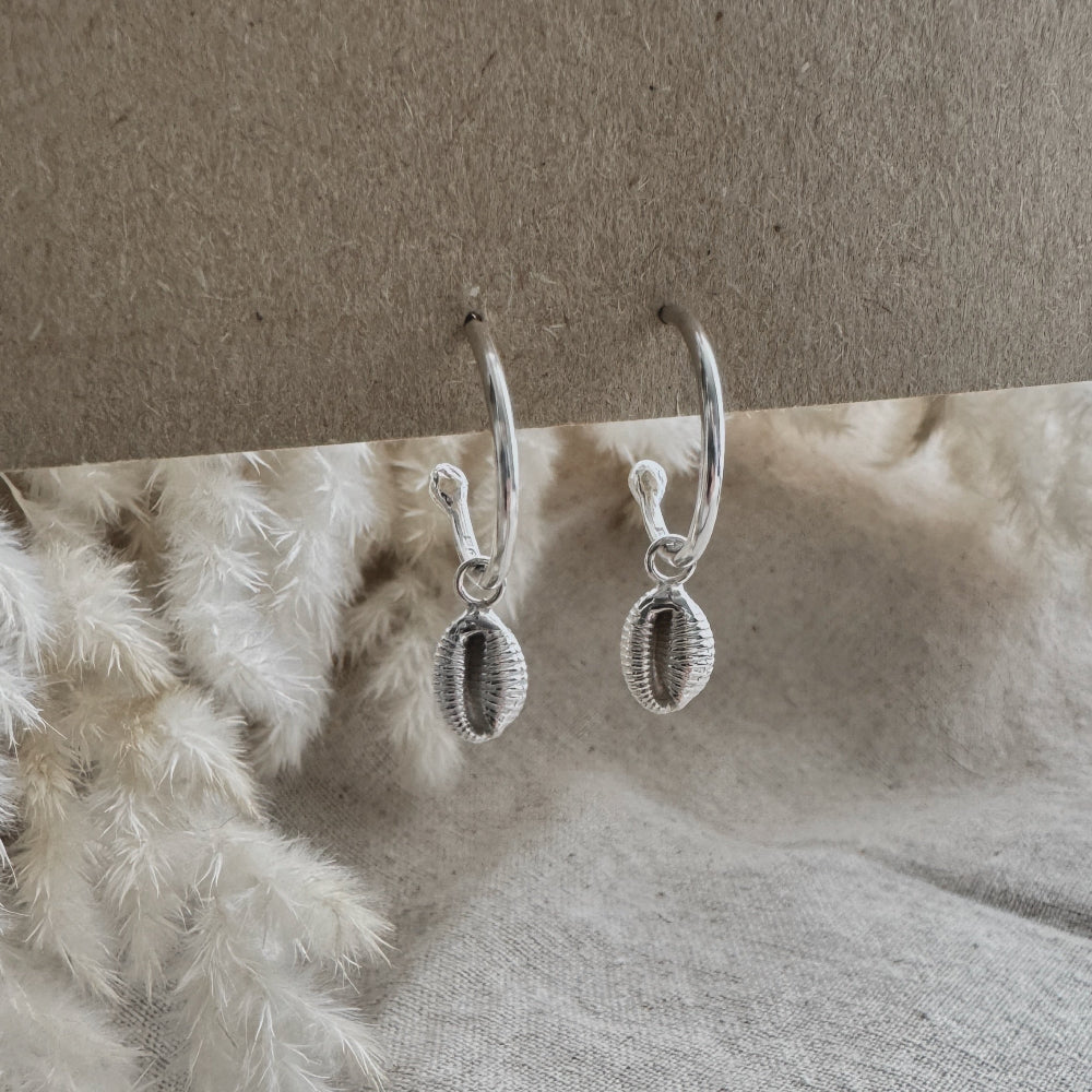 Silver hoop earrings with a cowrie shell charm on a textured surface with a neutral background