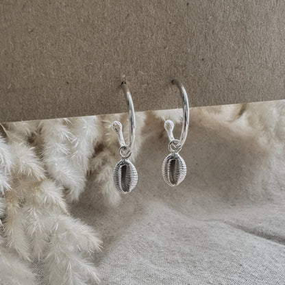 Silver hoop earrings with a cowrie shell charm on a textured surface with a neutral background