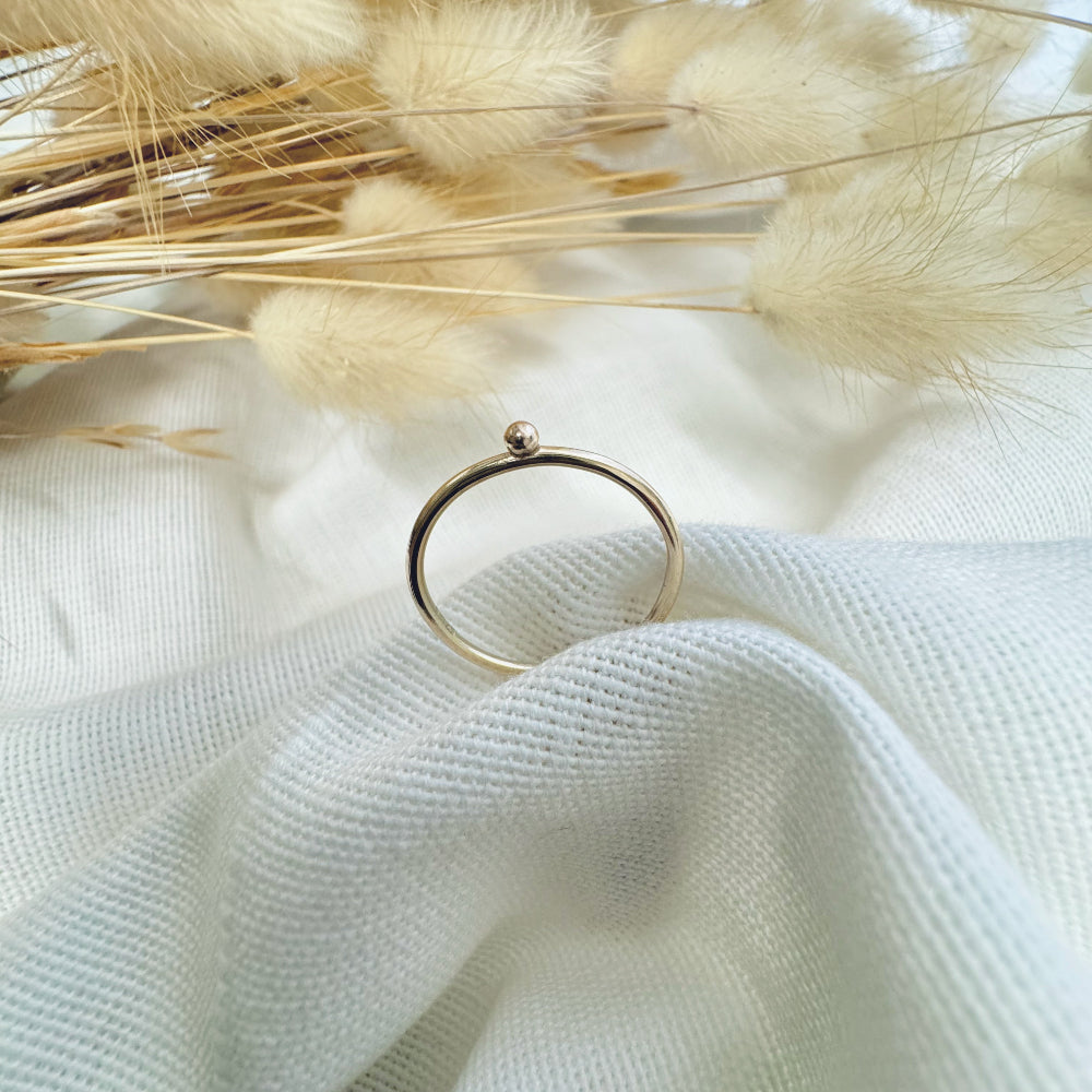A 9ct gold ball stacking ring with a small ball on top, placed on a white textured surface with dried grasses in the background