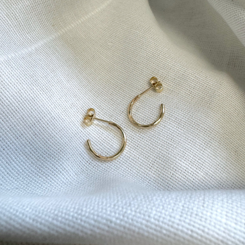 A pair of small hammered 9ct gold hoop earrings placed on a white textured surface 
