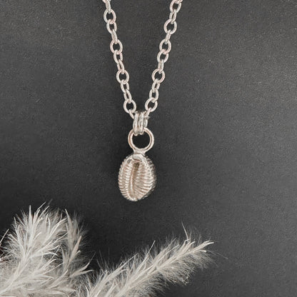 Silver cowrie shell necklace with a pendant on a grey surface with dried leaves.