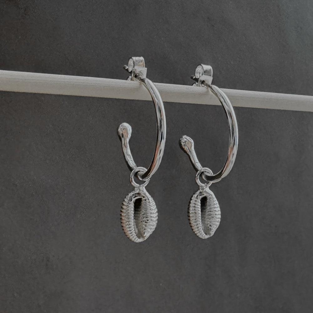 Pair of silver hoop earrings  with a cowrie shell charm on a dark gray background