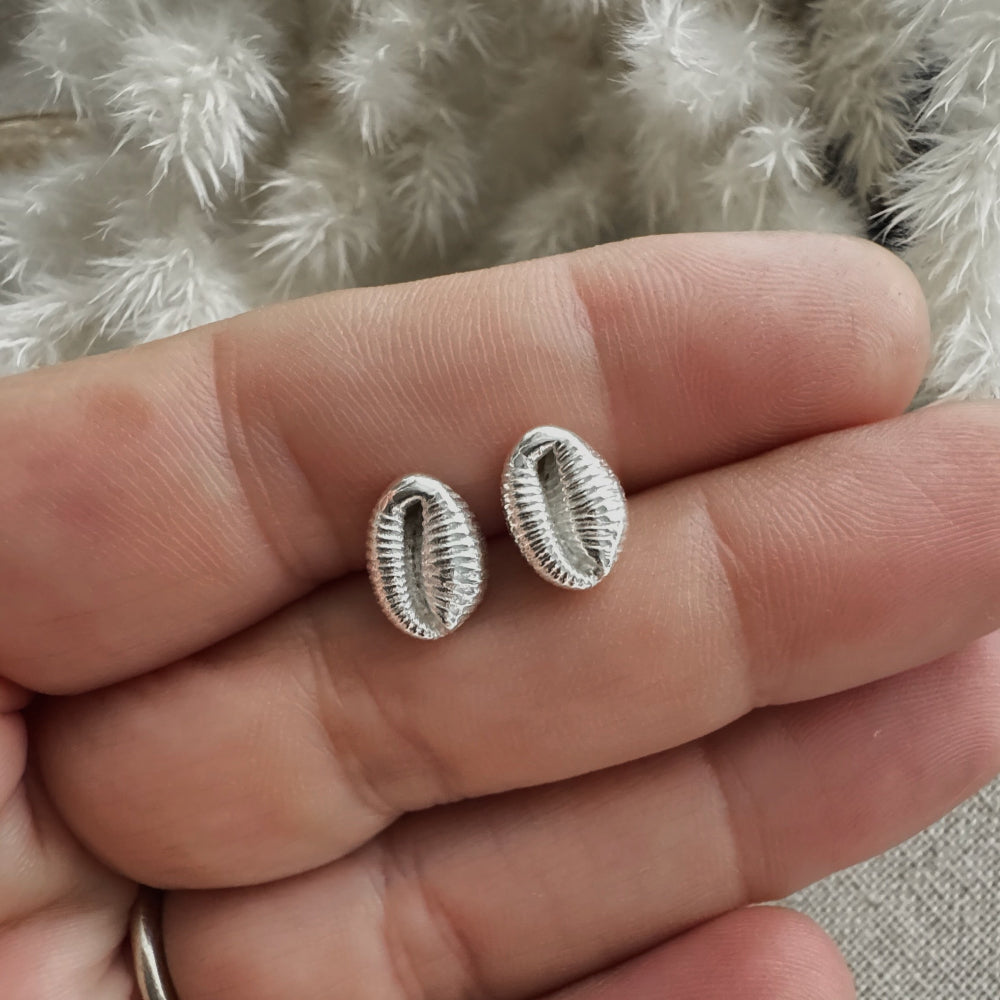 Silver Cowrie shell stud earrings held between two fingers of a hand against a textured fabric background
