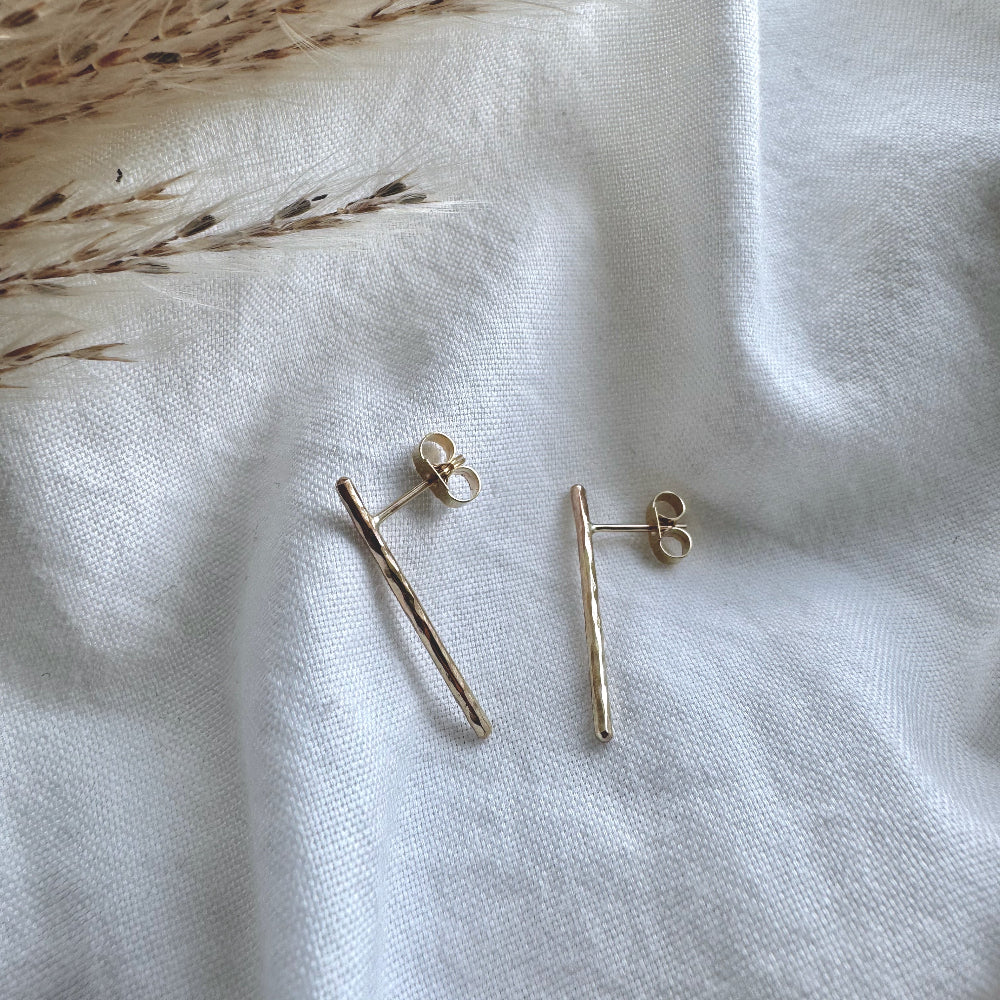 A pair of hammered gold bar earrings placed on a white fabric surface.