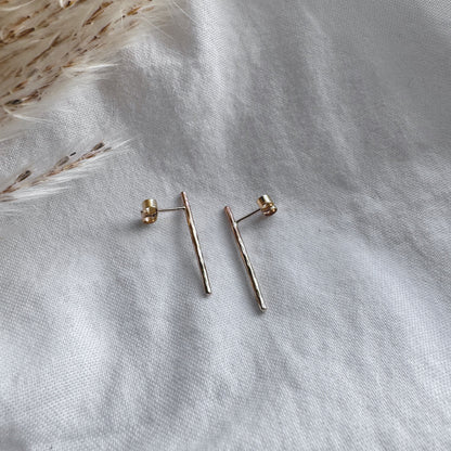 A pair of hammered gold bar earrings placed on a white fabric surface.