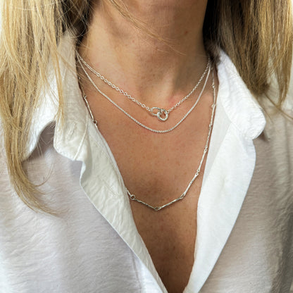 a model wearing a white shirt and 3 necklaces layer. the shortest has 2 silver links that are interlock,  the next is a fine silver chain and the longest one is a hammered bar chain necklace
