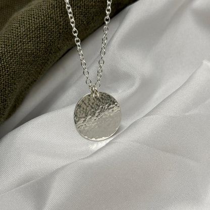 Silver hammered circle pendant hanging on a chain on a white fabric bacground