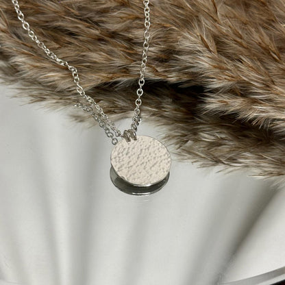 Round hammered silver pendant on a chain. On a mirrored background with dried grasses