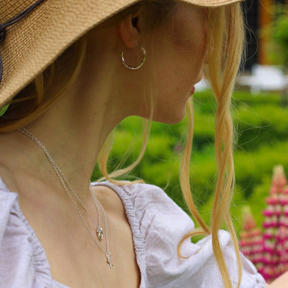 a lady wearing a straw hat and hammered hoops earrings with two dainty necklaces in a pale lilac top with a garden in the background
