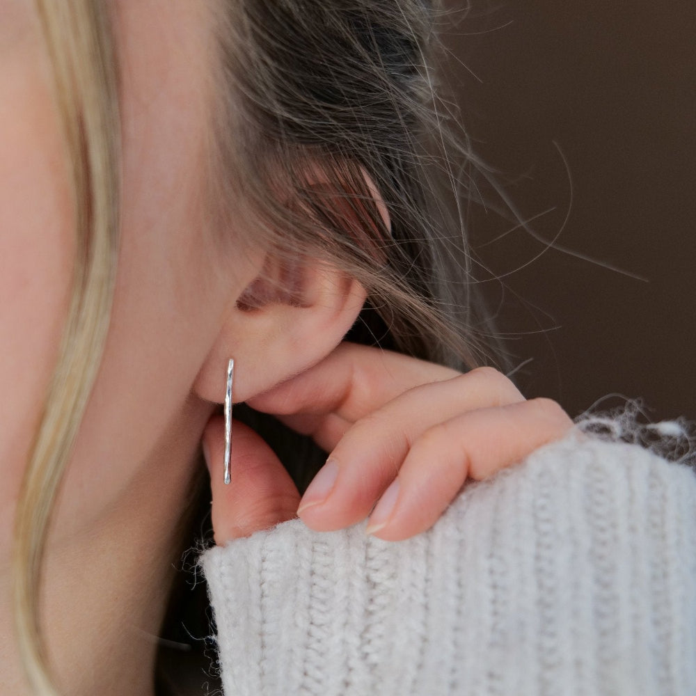 hammered silver bar earrings being worn in the ear of a lady and she's touching her ear wearing a cream jumper