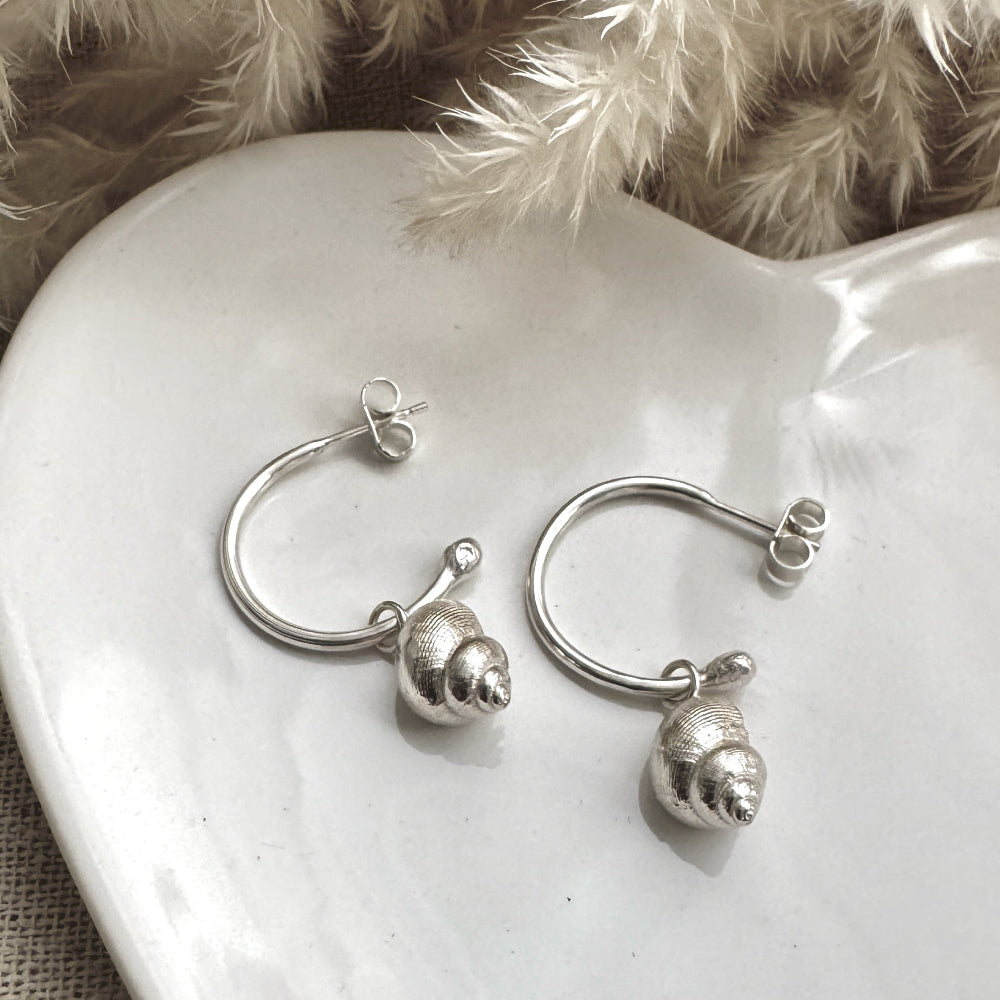 Pair of silver hoop earrings with a silver periwinkle charm on a white ceramic heart with a soft, neutral background.