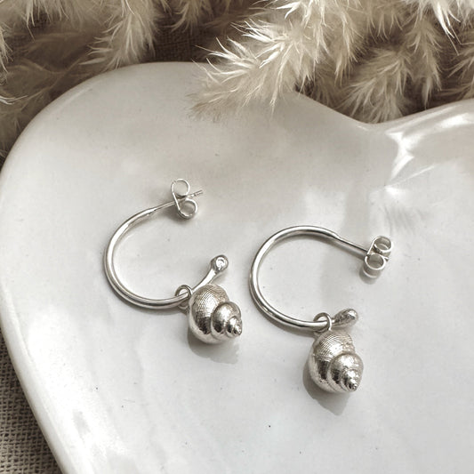 Pair of silver hoop earrings with a silver periwinkle charm on a white ceramic heart with a soft, neutral background.