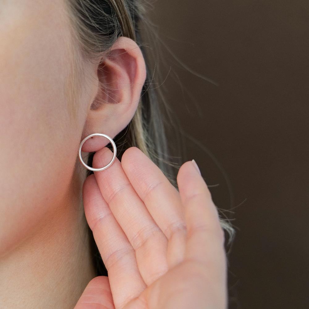 A close up of an ear with a lady wearing a large silver circle stud earring with her hand held up behind it