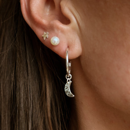 hammered silver Crescent moons as charms on silver hoop earrings being worn in the ear with a pearl stud and a star stud 