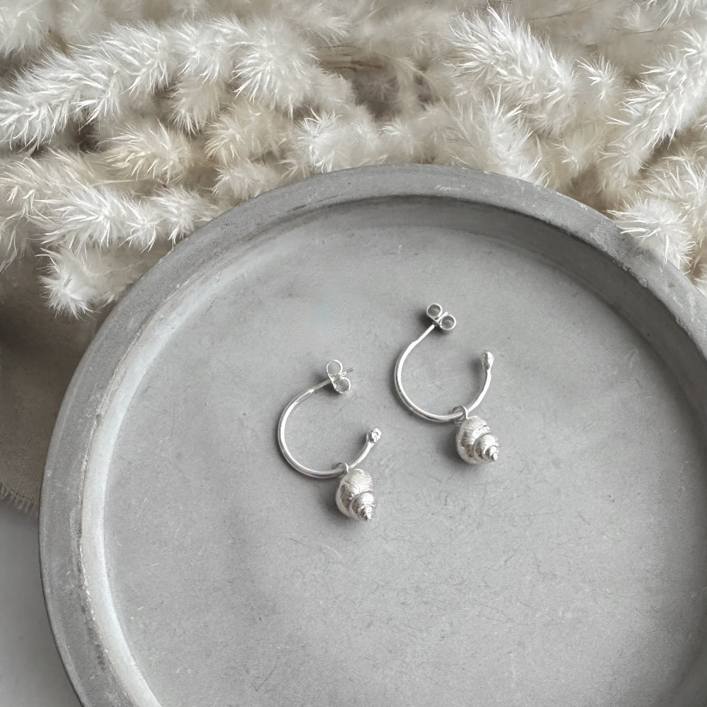 Silver hoop earrings with small silver periwinkle charmson a gray surface with a textured white background