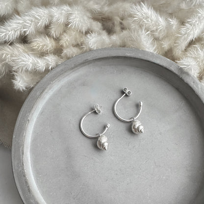 Silver hoop earrings with small silver periwinkle charmson a gray surface with a textured white background