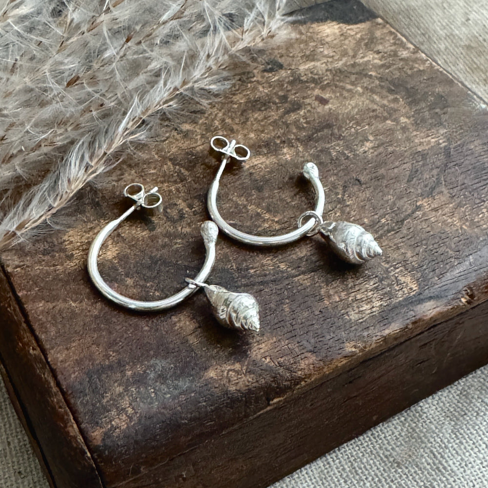 Silver hoop earrings with a whelk shell charm earrings on a rustic wooden surface with dried plants.
