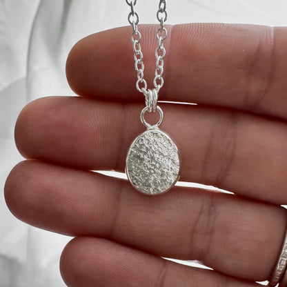Silver sand textured pendant necklace held between fingers against a neutral background