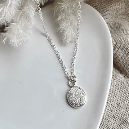 Silver necklace with a round textured pendant on a white plate with decorative feathers.