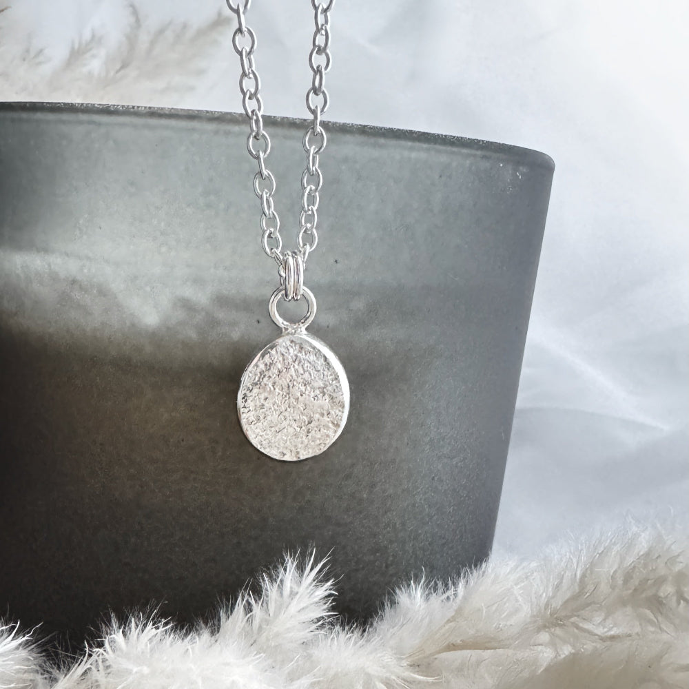 Silver necklace with a round, textured pendant in front of a grey glass on a white surface