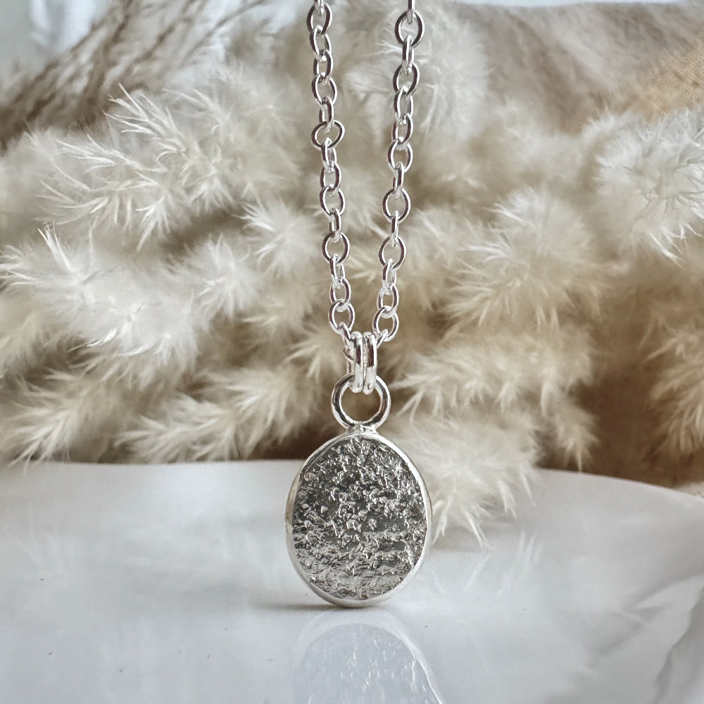 Silver necklace with a round textured pendant on a white surface with a fluffy background