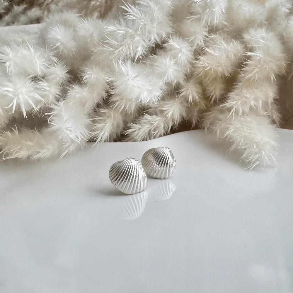 Silver cockle shell earrings on a white surface with a textured beige fabric background with dried grasses