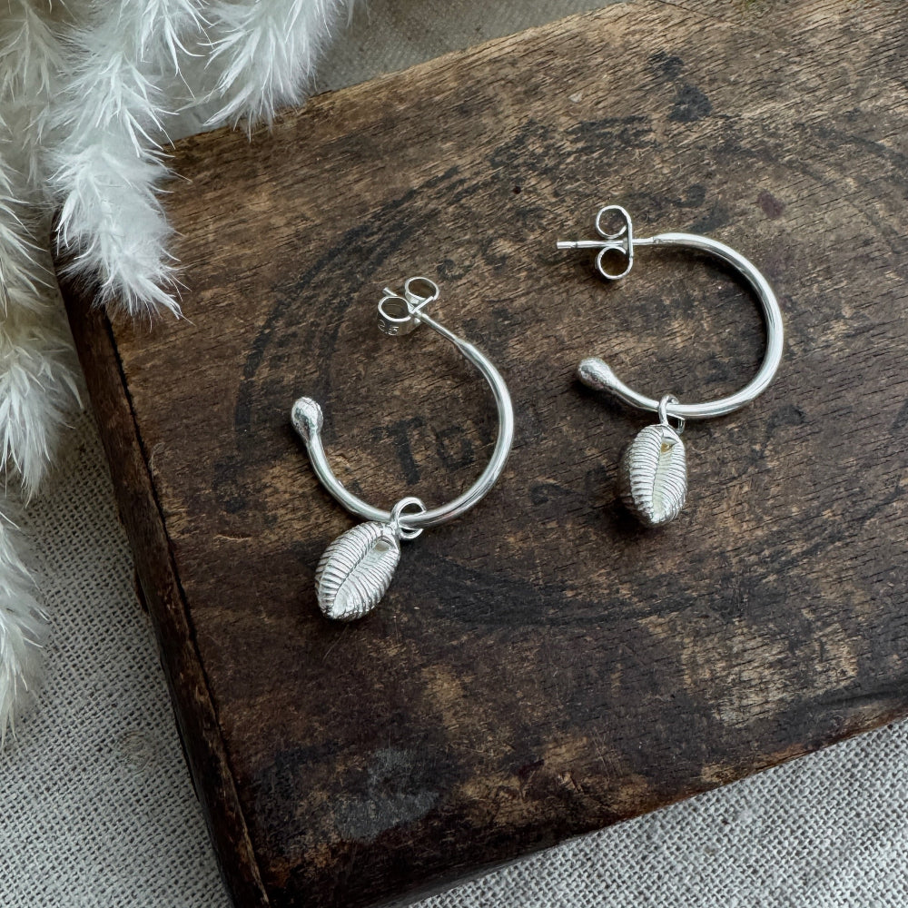 Open Silver hoop earrings with cowrie shell charms on a wooden surface.