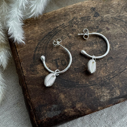 Open Silver hoop earrings with cowrie shell charms on a wooden surface.