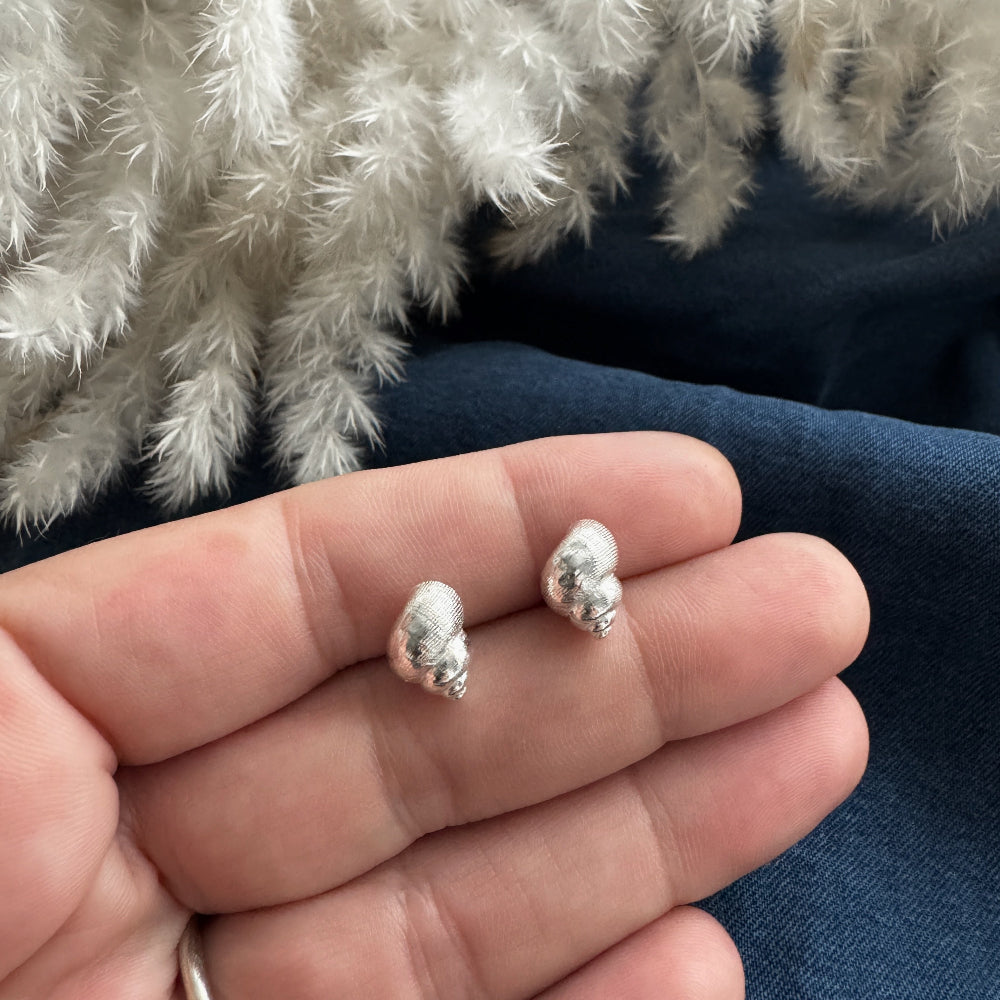 Silver periwinkle shell earrings held between fingers against a textured fabric background