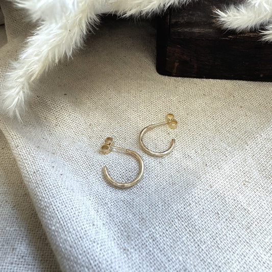 A pair of small hammered 9ct gold hoop earrings placed on a textured surface with a white furry object in the background.