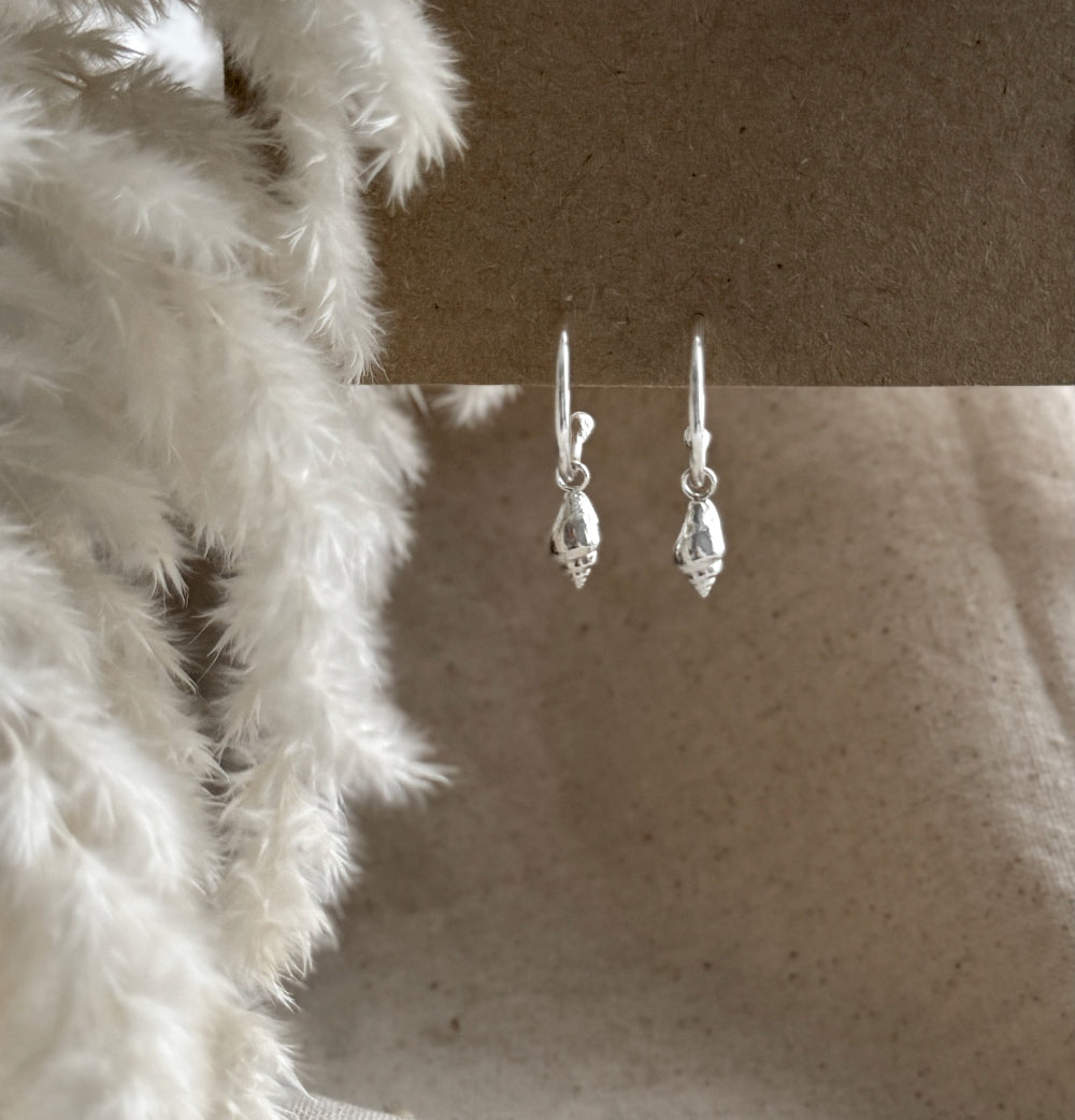Silver hoop earrings with a whelk shell charm on a textured surface with a soft white background