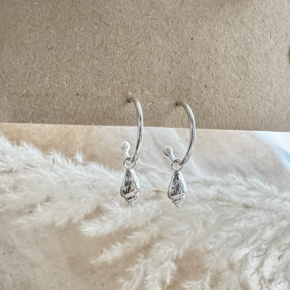 Silver hoop earrings with a whelk charms on a textured beige surface.