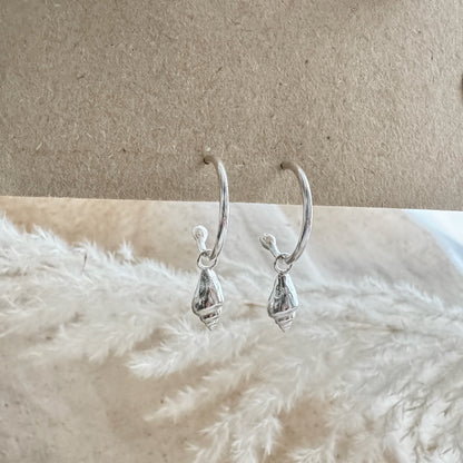 Silver hoop earrings with a whelk charms on a textured beige surface.