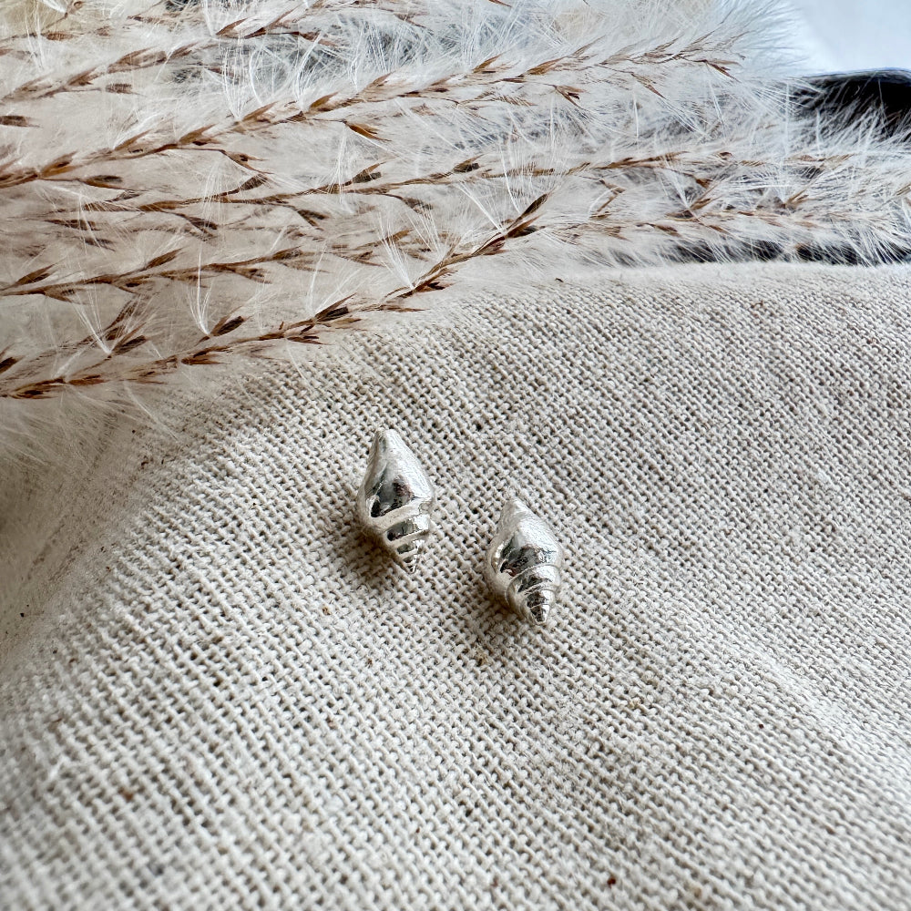 Silver whelk shell earrings on a textured fabric surface with dried grasses.