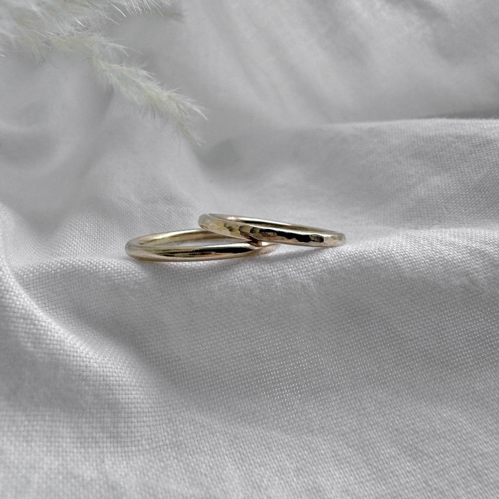 two 9ct gold rings the layed down overlapping each other. The bottom one is plain the top one hammered and they are placed on a white fabric with a feather-like texture in the background.