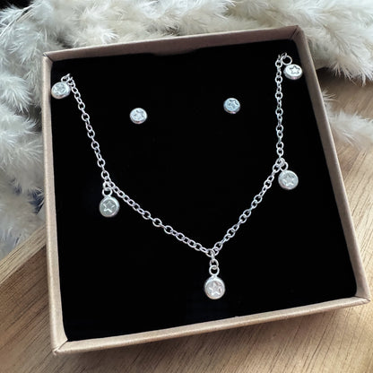 Silver necklace and earrings set in a box on a wooden surface with a fluffy white foliage in the background.