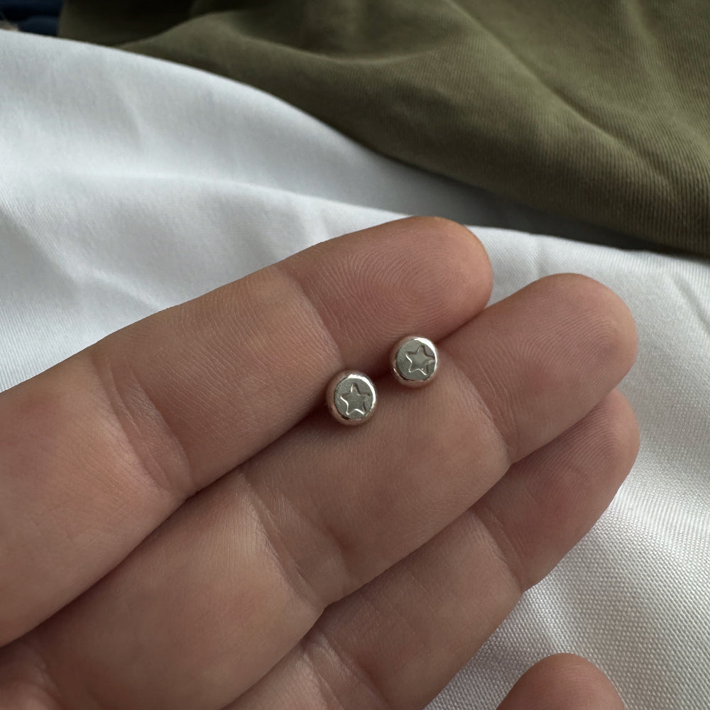 Small silver earrings with a star stamped into them held between fingers on a neutral background