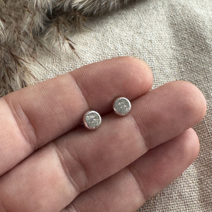 Two stud earrings held between fingers against a textured fabric background