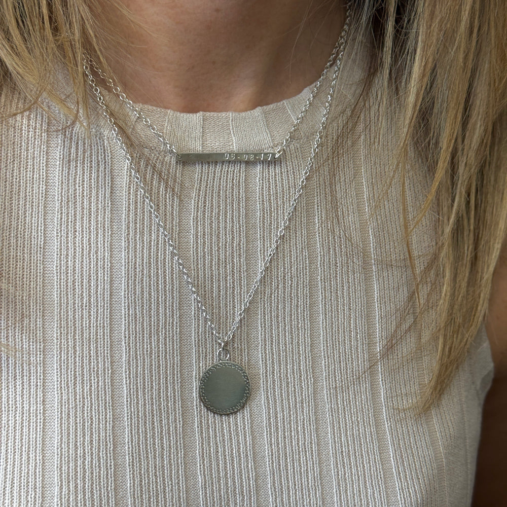 a lady wearing two necklace on over a cream top. the shortest on is a personalised bar necklace with numbers and the longest one is a silver circle with a textured edge