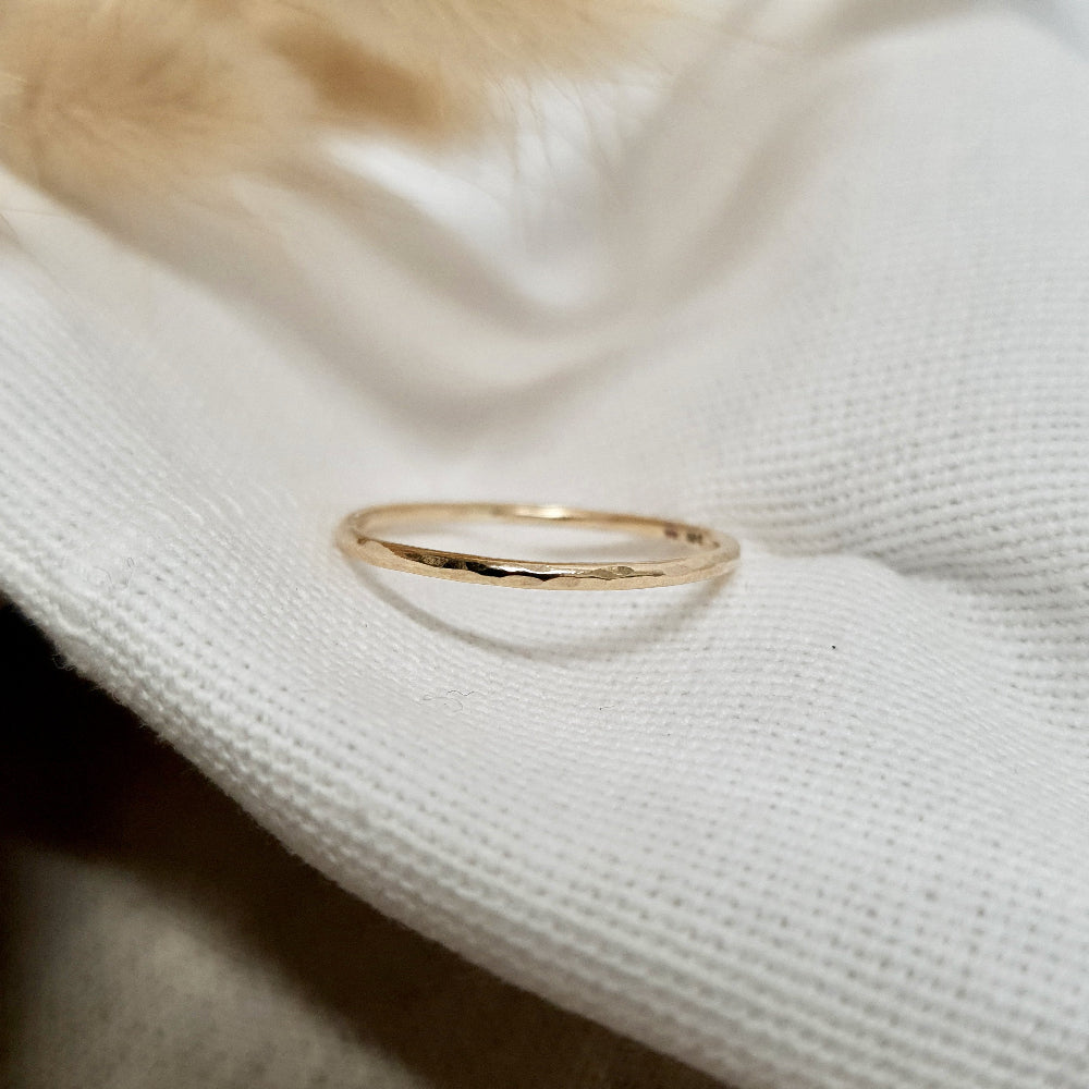 A single hammered gold stacking ring displayed against a neutral background with dried flowers to the side.