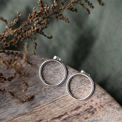A pair of large silver circle stud earrings placed on a wooden surface with a blurred background of plant leaves.