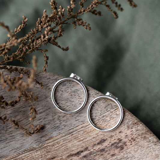 A pair of large silver circle stud earrings placed on a wooden surface with a blurred background of plant leaves.