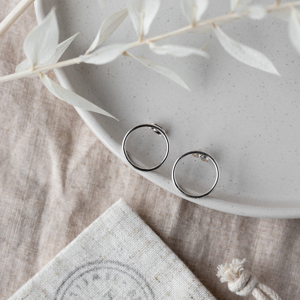 A pair of large silver circle stud earrings placed on a ceramic plate with a blurred background of plant leaves.