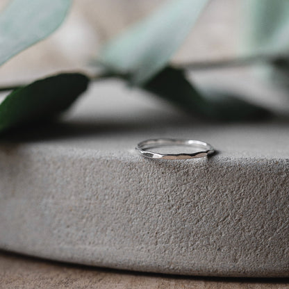 A simple, elegant silver stacking ring with a hammered finish, displayed on a grey background with dried plants behind it.