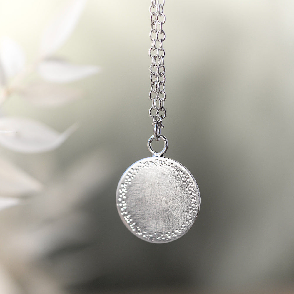 a silver pendant with a textured edge hanging on a delicate chain on a blurred background